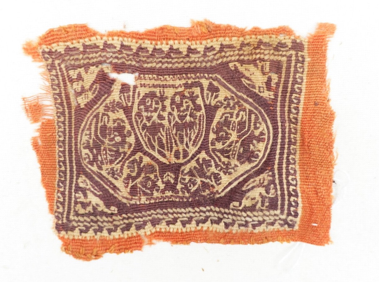 Fragment of cloth with a rectangle enclosing an oval with stylised paired human figures and foliage surrounded by very stylised animals (rabbit?) in dark purple wool and linen, on an unbleached linen ground and part of a red woollen ground. Hand spun and woven (dovetail tapestry, weft-faced). Coptic, mid-first millennium. Dool family donation (TRC 2023.0071).