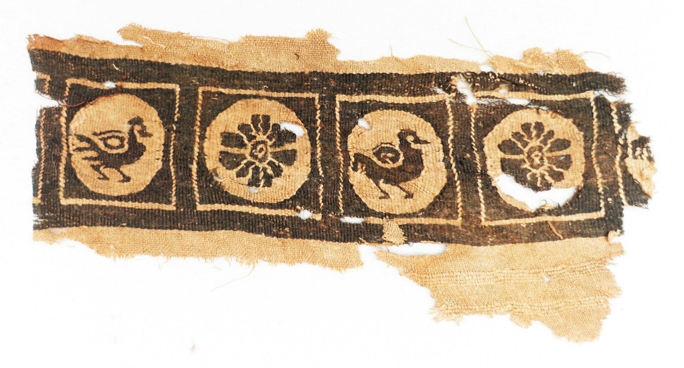 Fragment of cloth with a broad band with individual squares enclosing stylised birds and rosettes in black wool on an unbleached linen ground. Hand spun and woven (tapestry, plain ground). Coptic, mid-first millennium. Dool family donation (TRC 2023.0075).