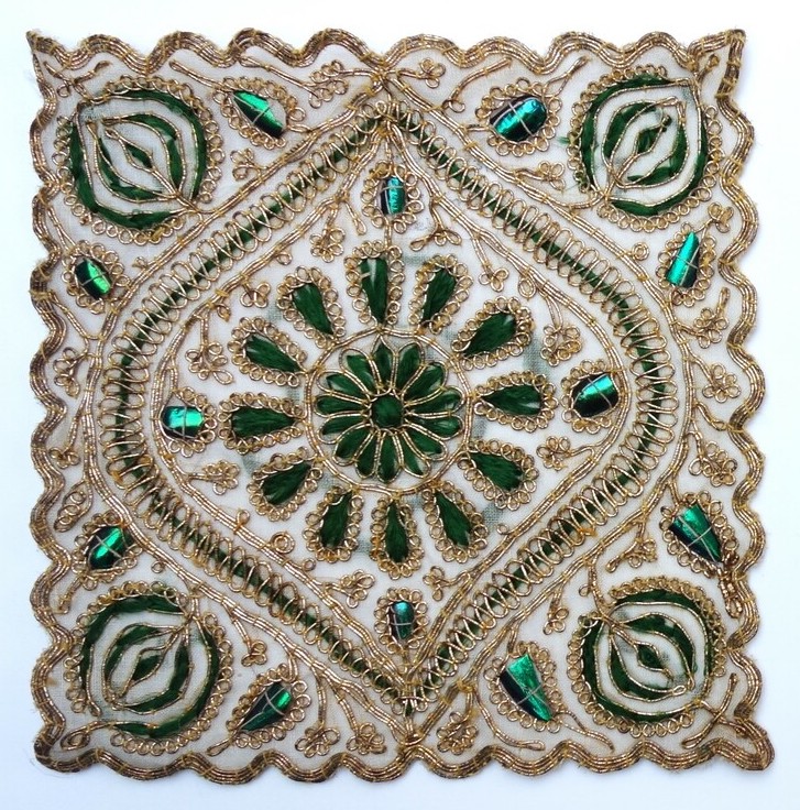 A square mat from India, 1920s, decorated with embroidered rosettes, ovals, ogees, and circles worked in green satin stitch, pieces of beetle wing casings and gold-coloured metal thread worked in various manners (TRC 2025.0331a).