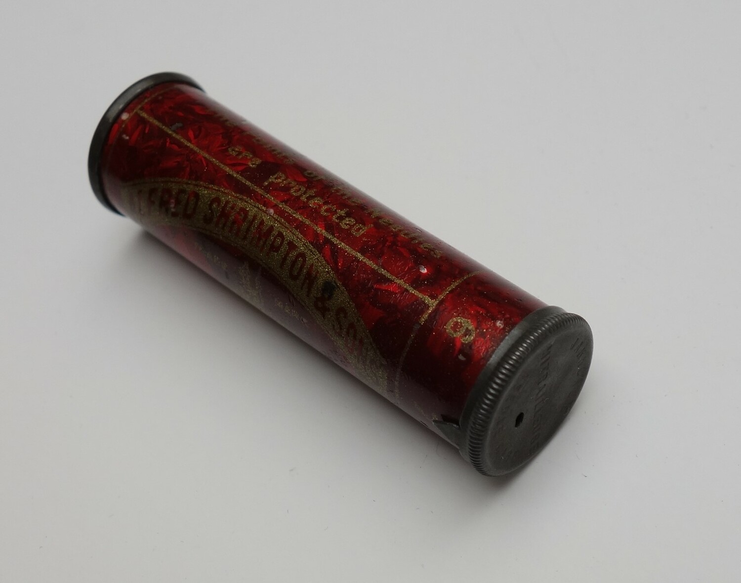 Round red metal case containing needles. The packaging has the name ALFRED SHRIMPTON & SONS, REDDITCH. The circular lid has a small hole for the needles. UK, 1890-1900. TRC 2025.0672).
